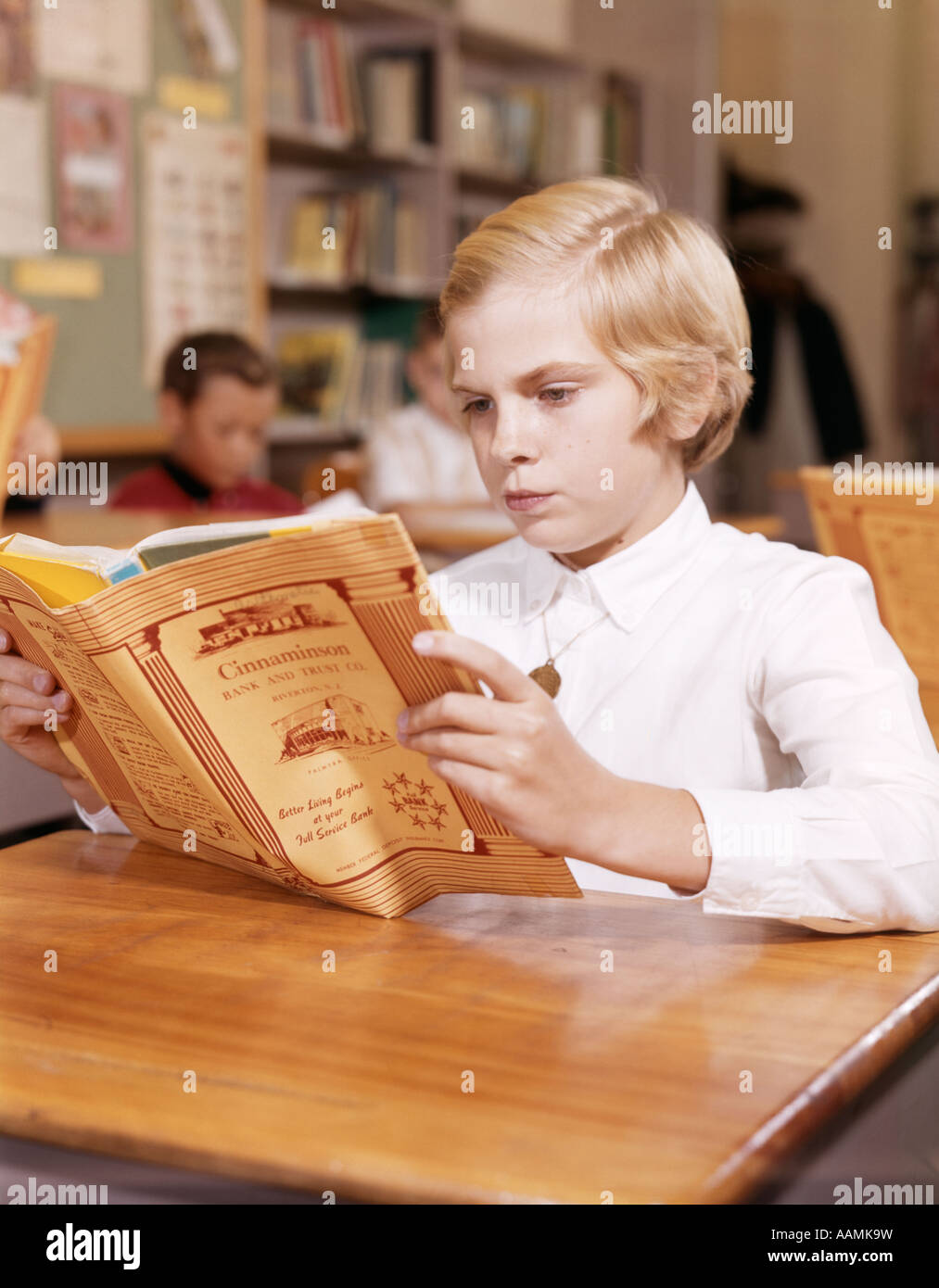 1960s BLONDE GIRL READING BOOK AT DESK IN CLASSROOM Stock Photo