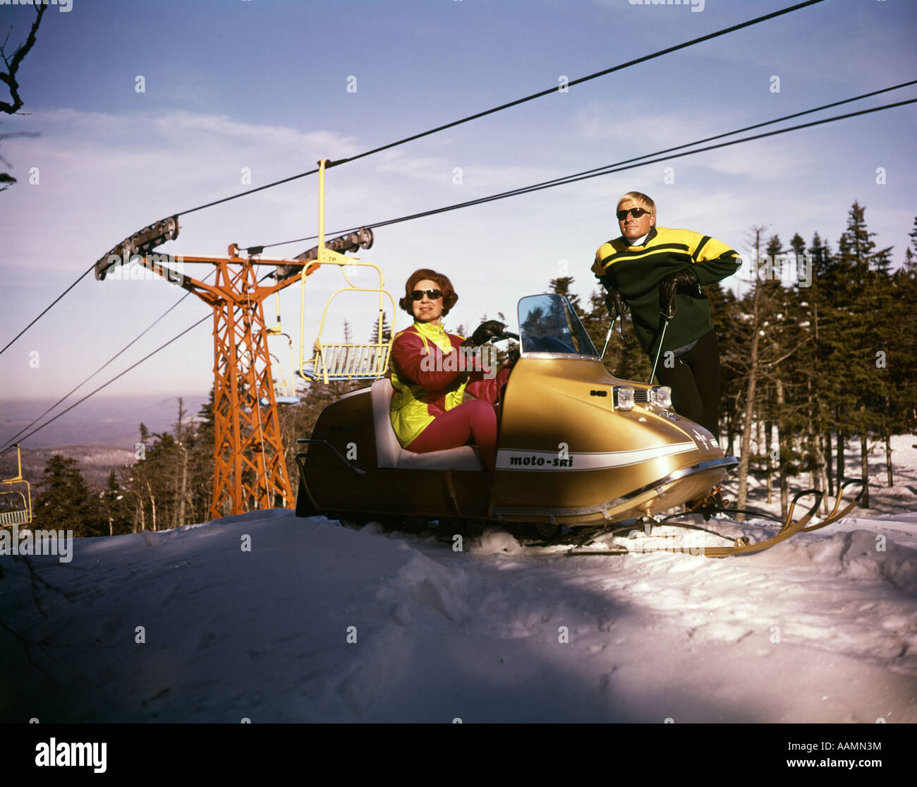 1960s MAN ON SKIS TALKING TO WOMAN IN SNOWMOBILE NEAR SKI LIFT IN MOUNTAINS LIFESTYLE VACATION HOBBY Stock Photo