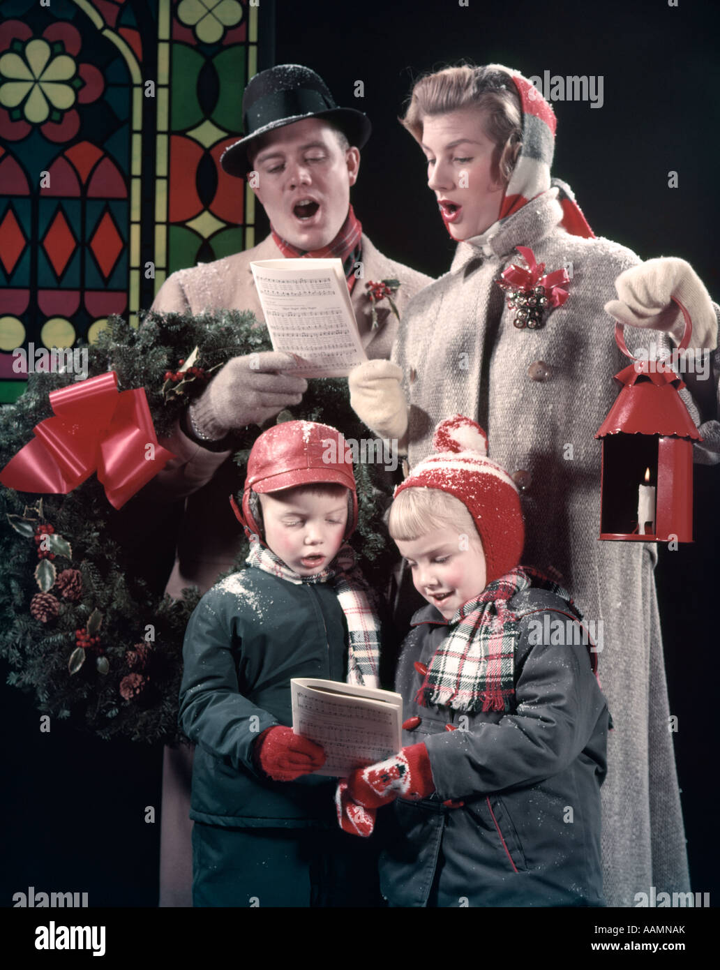 1950s FAMILY 4 SINGING CAROLS DAD MAN HAS WREATH OVER ARM MOM WOMAN HOLDS RED LANTERN BOY GIRL MUSIC SONG SNOW OUTDOOR Stock Photo