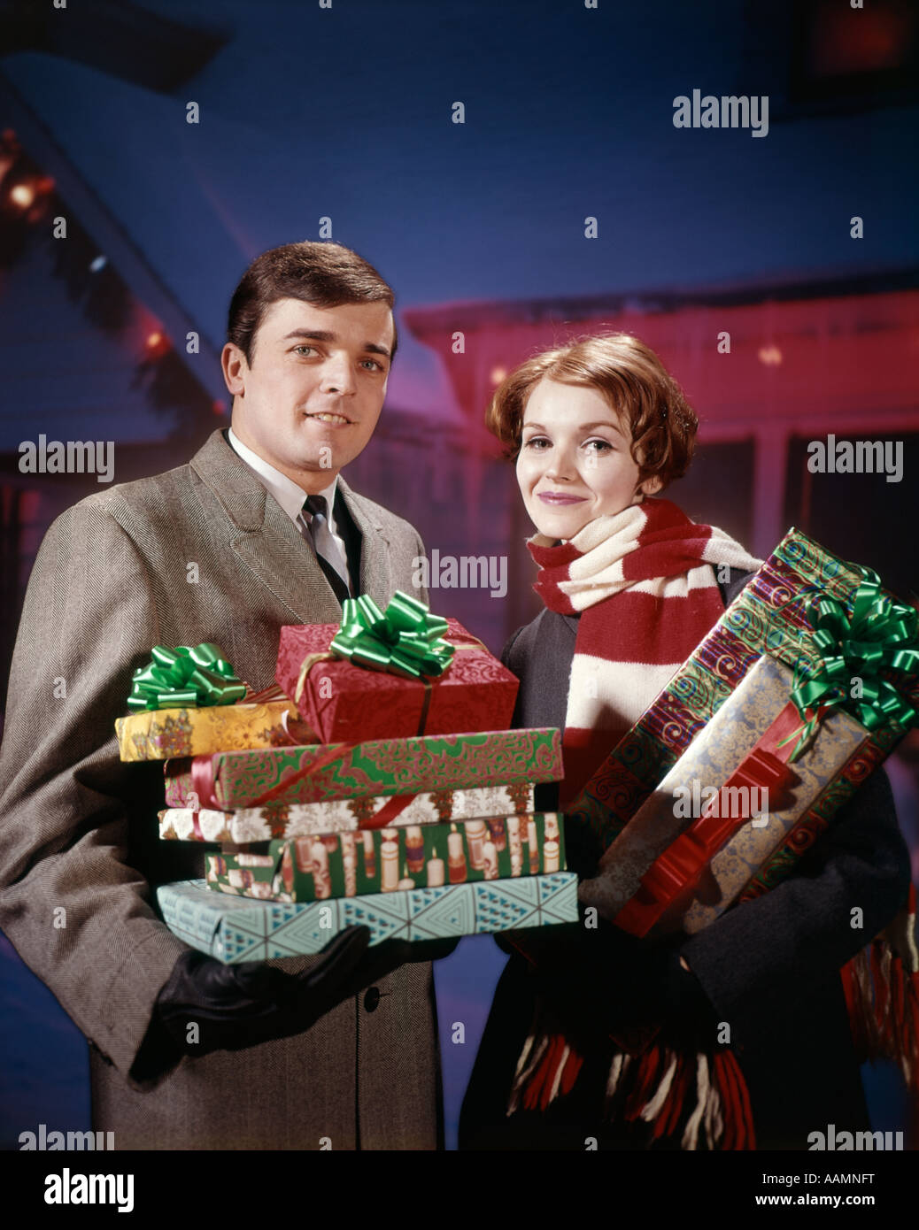 1960 1960s 1970 1970s COUPLE MAN WOMAN HOLDING STOCK PRESENTS GIFTS IN FRONT OF HOUSE DECORATED WITH LIGHTS STUDIO RETRO Stock Photo
