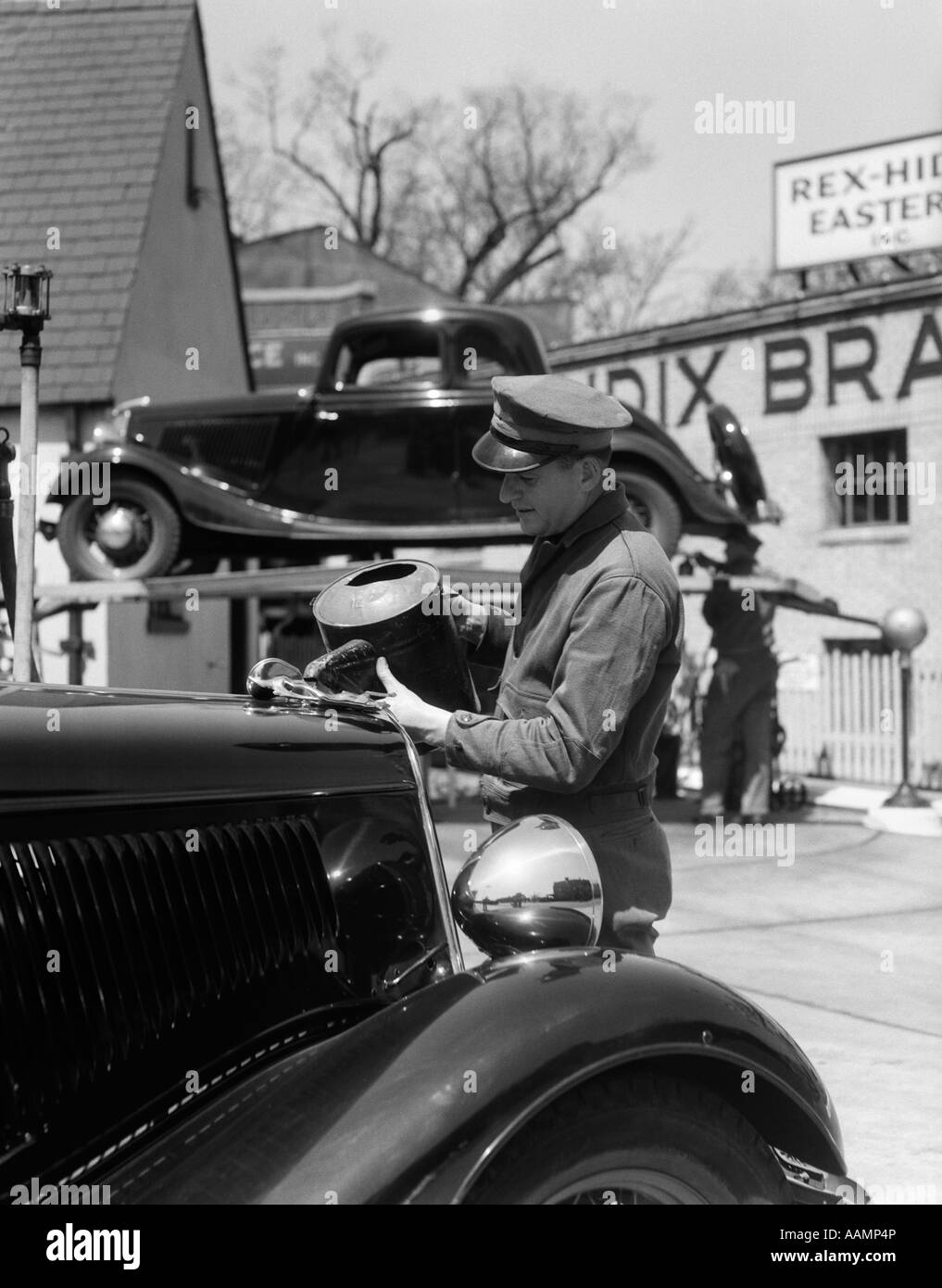 1930s SERVICE STATION ATTENDANT IN CAP & COVERALLS POURING WATER FROM SPOUTED CAN INTO AUTOMOBILE RADIATOR Stock Photo