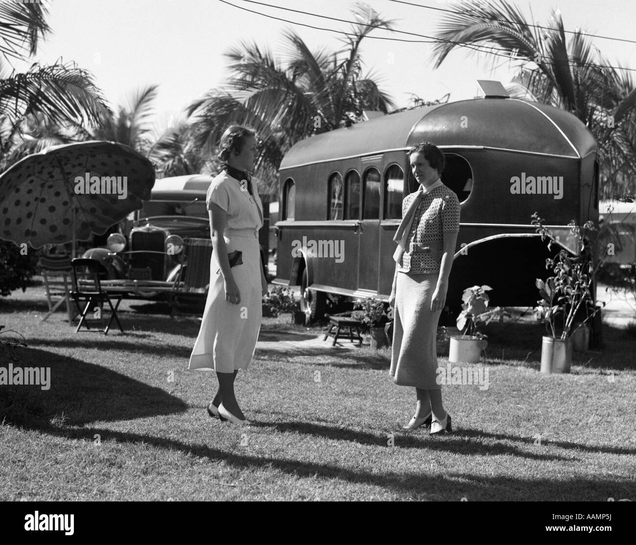 1930s 2 WOMEN STANDING TALKING ON TROPICAL LAWN IN FLORIDA TRAILER PARK WITH A CAR UMBRELLA & CHAIRS Stock Photo
