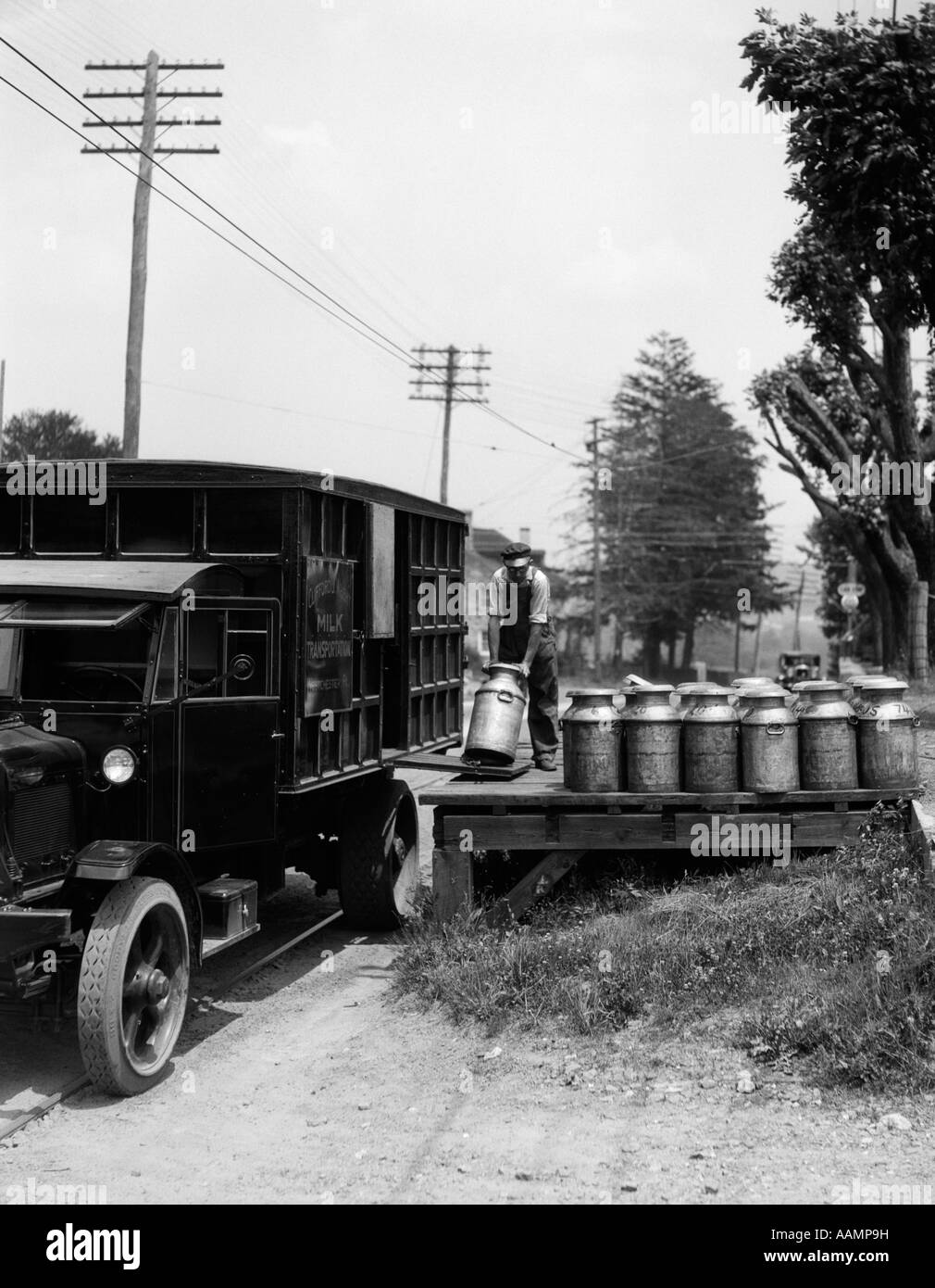 1920s DELIVERY MAN UNLOADING MILK CANS FROM LARGE TRUCK ONTO WOODEN PLATFORM ALONG SIDE OF ROAD Stock Photo
