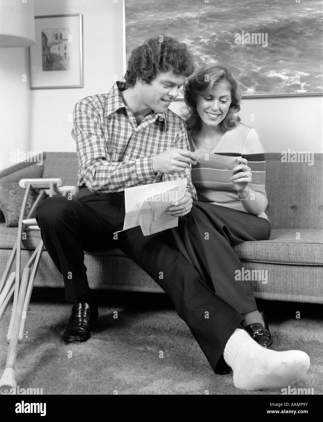 1970s MAN AND WOMAN BROKEN FOOT ON STOOL CRUTCHES SMILING AT INSURANCE CHECK Stock Photo