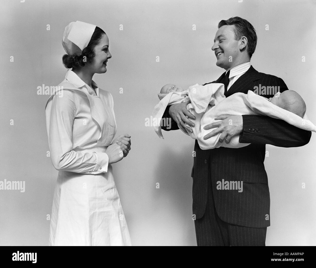 1930s 1940s SMILING WOMAN NURSE TALKING WITH MAN PROUD NEW FATHER HOLDING NEWBORN TWIN BABIES Stock Photo