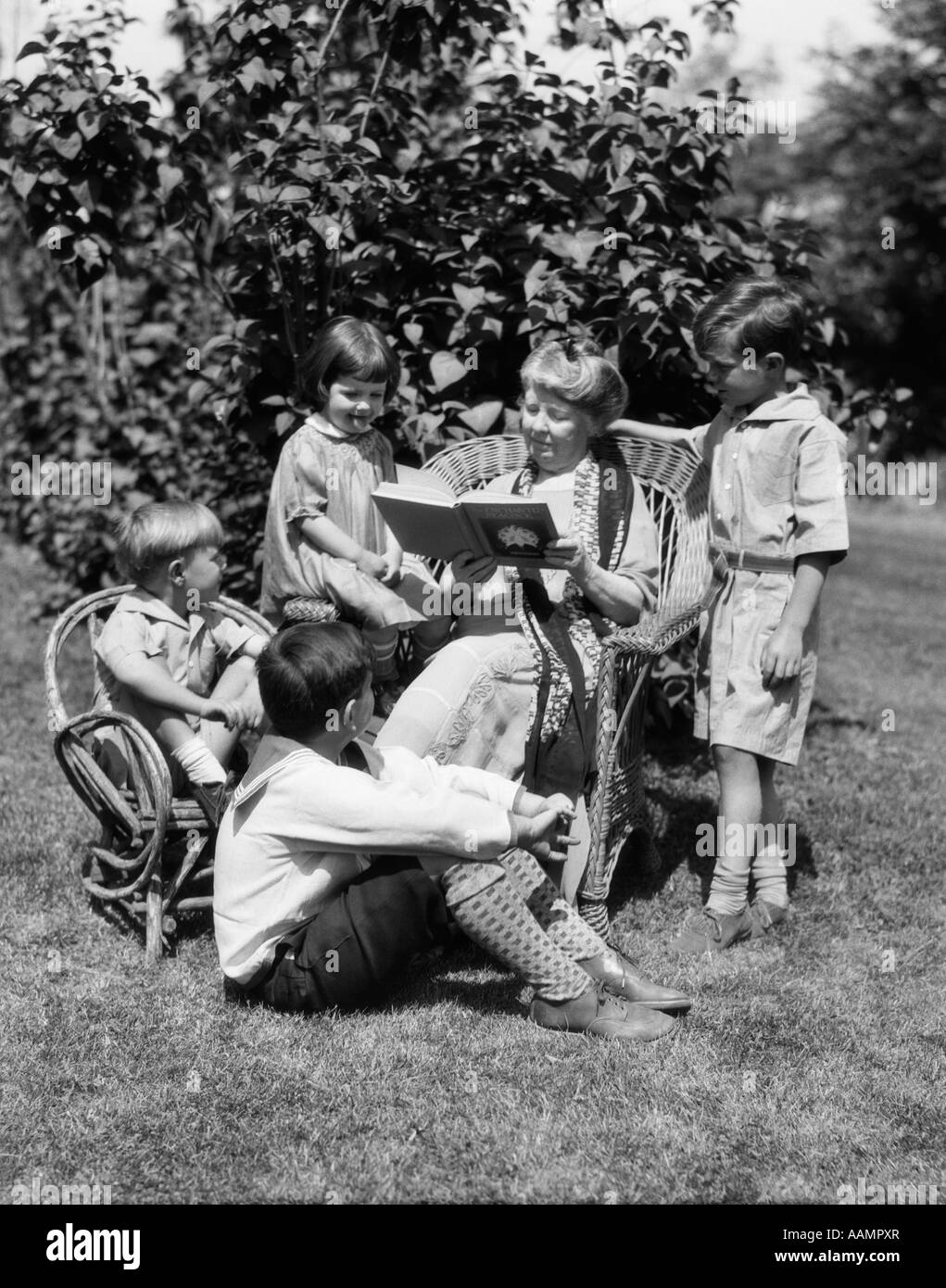 1920s OLDER WOMAN GRANDMOTHER SITTING IN WICKER CHAIR READING BOOK TO FOUR BOYS GIRLS GRANDCHILDREN Stock Photo