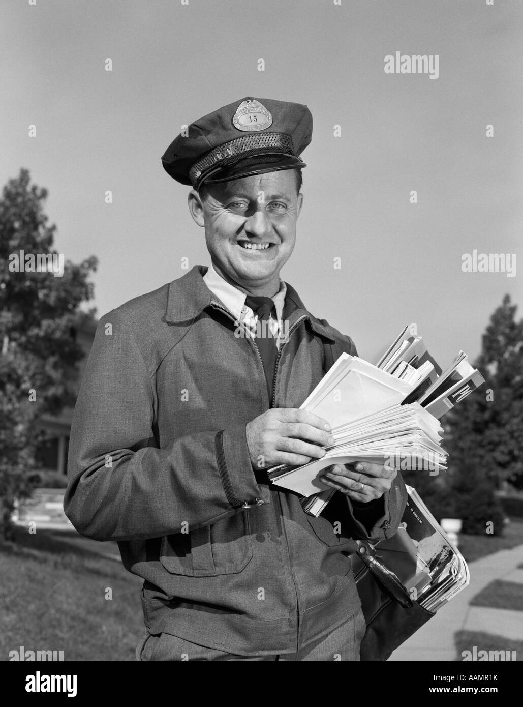 1960s SMILING MAILMAN OUTDOORS IN SUBURBAN NEIGHBORHOOD HOLDING LETTERS MAILBAG Stock Photo
