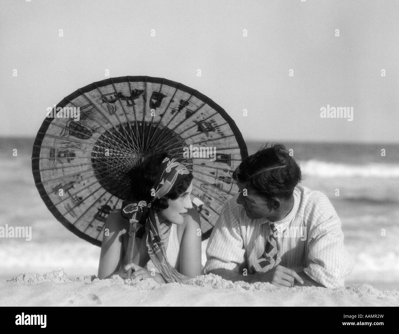 1920s ROMANTIC COUPLE MAN WOMAN LOOKING AT ONE ANOTHER LYING FACE TO FACE UNDER ORIENTAL PARASOL ON SANDY BEACH Stock Photo