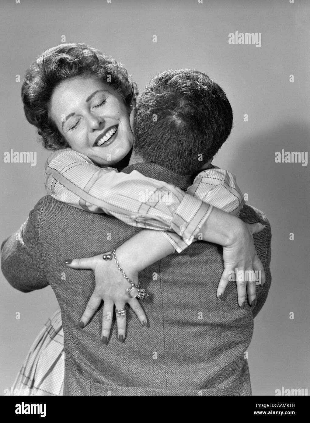 1950s COUPLE HAPPY SMILING WOMAN FACING FORWARD HANDS DRAPED OVER MANS BACK HUGGING EMBRACING THRILLED LOVE Stock Photo