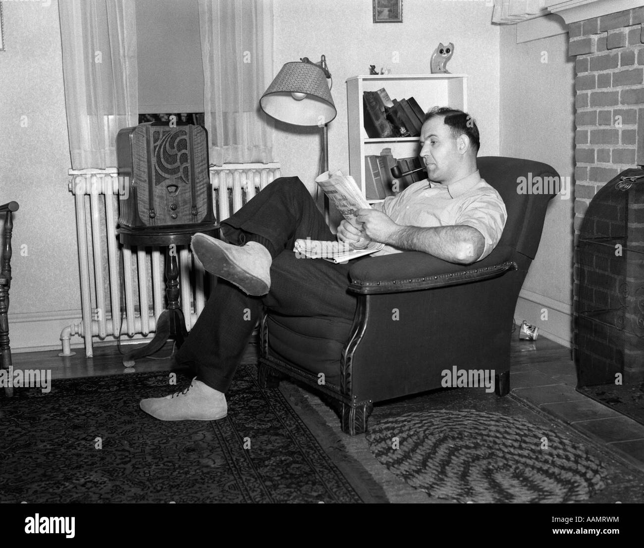 1950s MAN SITTING IN CHAIR IN LIVING ROOM SMOKING PIPE READING PAPER LISTENING TO RADIO Stock Photo
