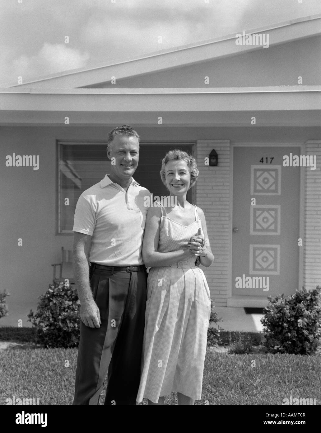 1950s SMILING OLDER MAN WOMAN SENIOR CITIZEN STANDING TOGETHER IN RETIREMENT HOME FRONT YARD LOOKING AT CAMERA Stock Photo