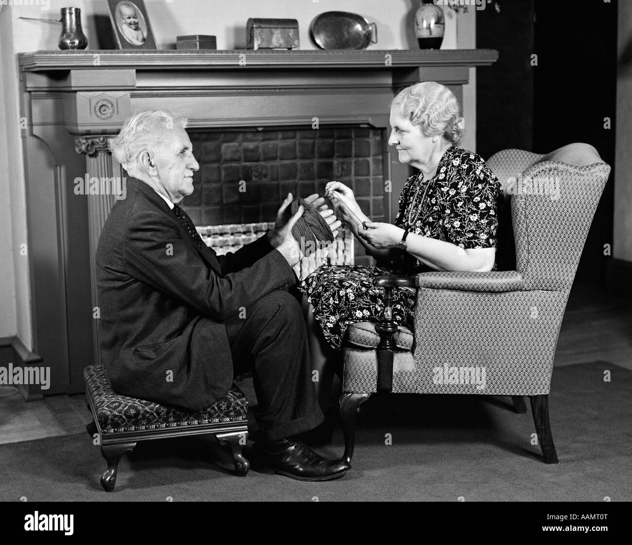1940s SIDE VIEW OF ELDERLY MAN ON STOOL HOLDING HANDS OUT FOR WIFE IN CHAIR OPPOSITE HIM TO WRAP YARN FOR KNITTING Stock Photo