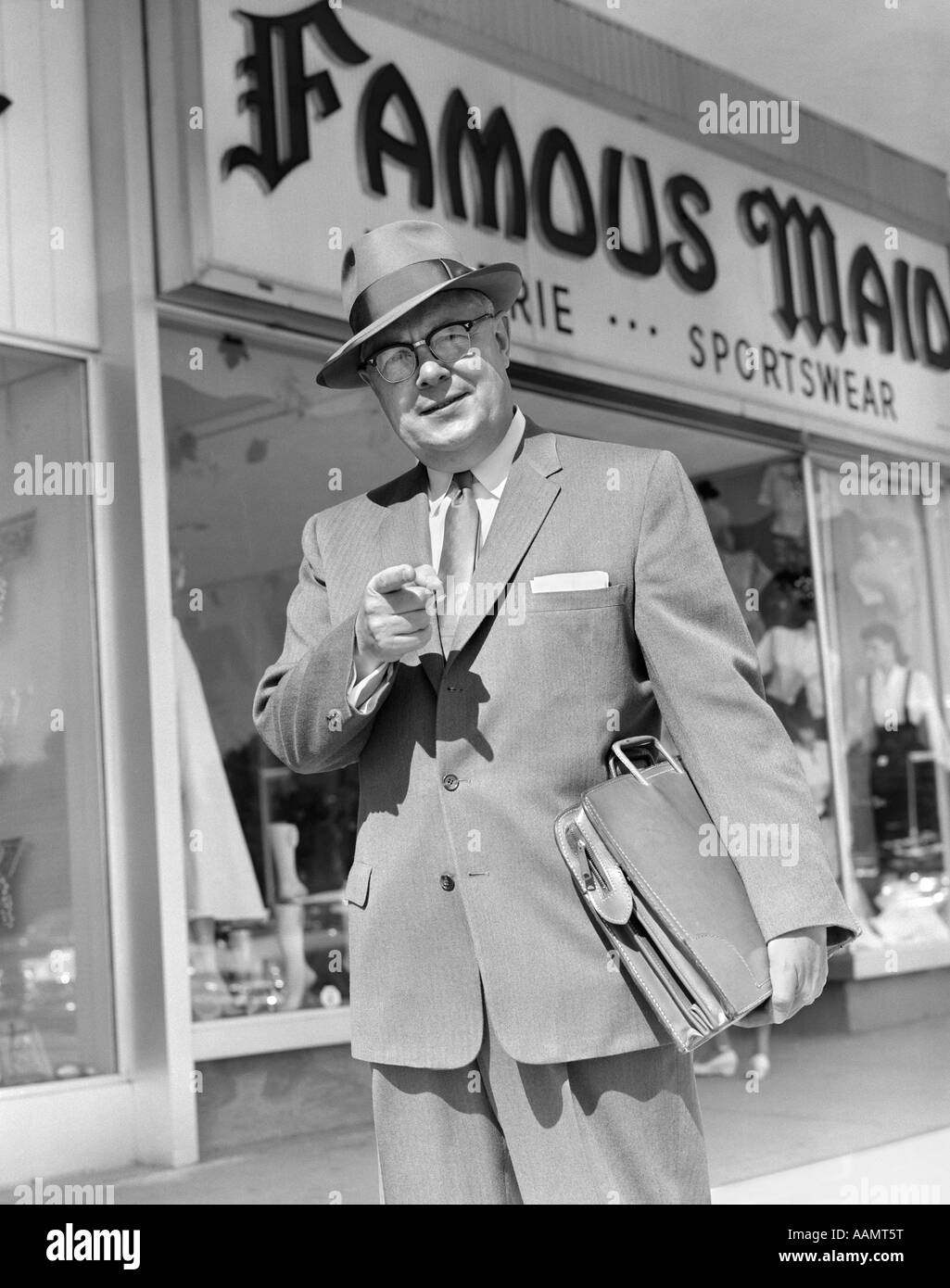 1950s MAN POINTING TO CAMERA HOLDING BRIEFCASE UNDER ARM IN FRONT OF SPORTS WEAR STORE SELL SUIT TIE HAT BUSINESSMAN Stock Photo