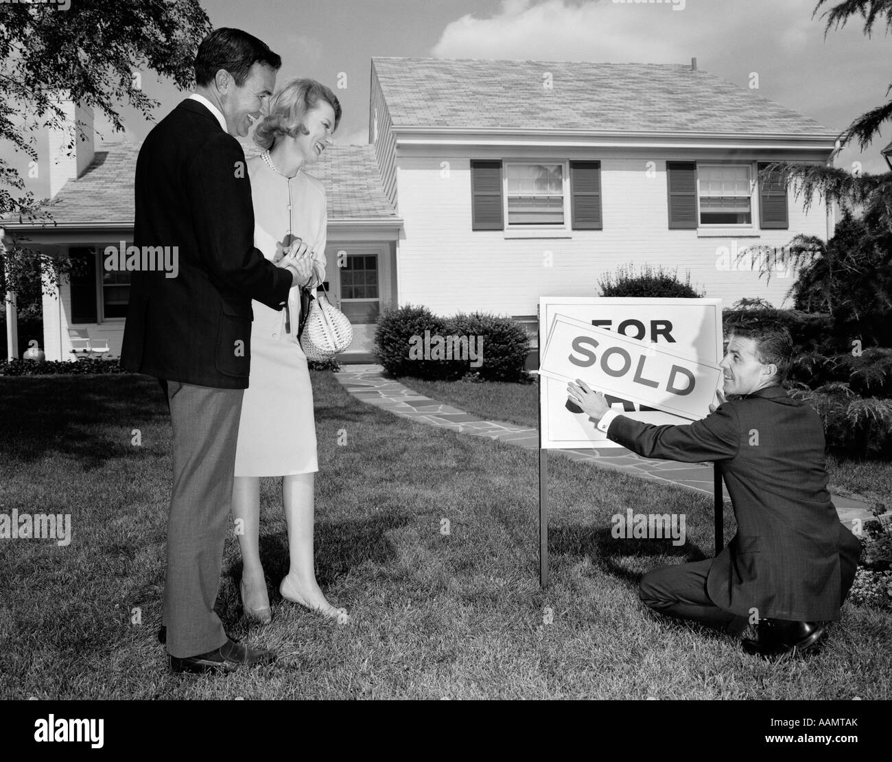 1960s SMILING COUPLE STANDING ON FRONT LAWN OF NEW HOUSE LOOKING DOWN AT REALTOR PUTTING UP SOLD SIGN Stock Photo
