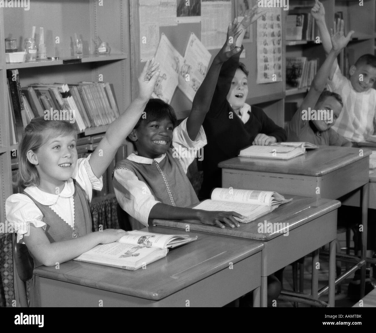 1960s GROUP OF GRADE SCHOOL CHILDREN AT ROW OF DESKS EAGERLY RAISING HANDS Stock Photo