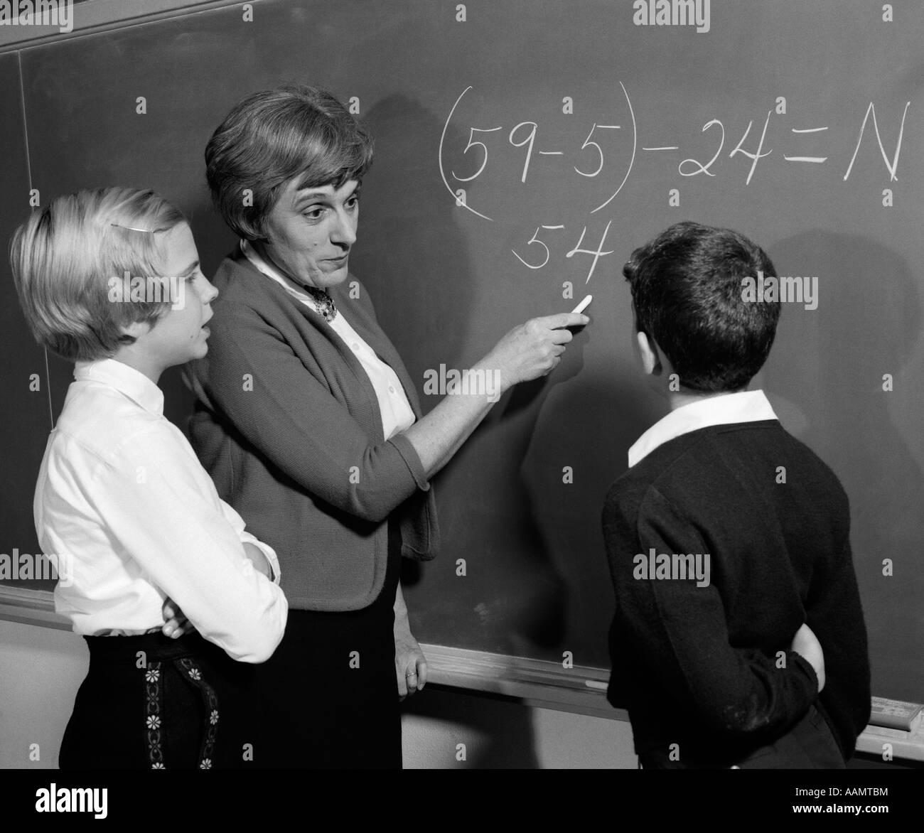 1960s TEACHER AT BLACKBOARD WITH MALE & FEMALE STUDENT EXPLAINING MATH PROBLEM Stock Photo