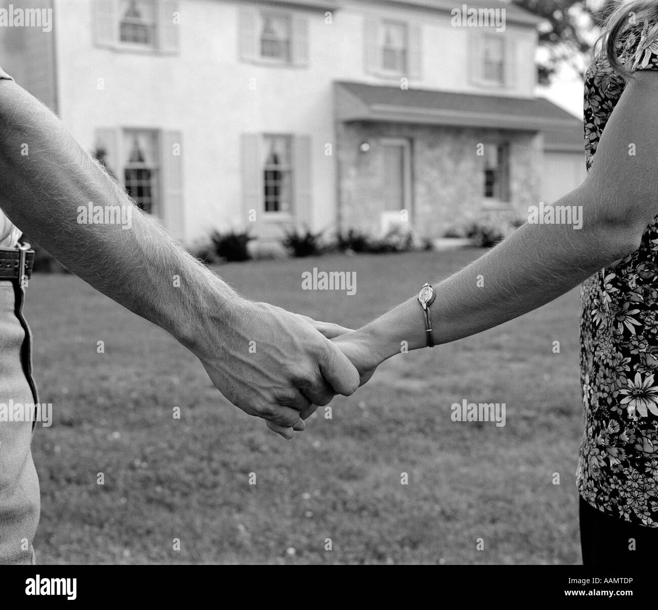 1970s CLOSE-UP OF COUPLE HOLDING HANDS WITH NEW HOME IN BACKGROUND Stock Photo