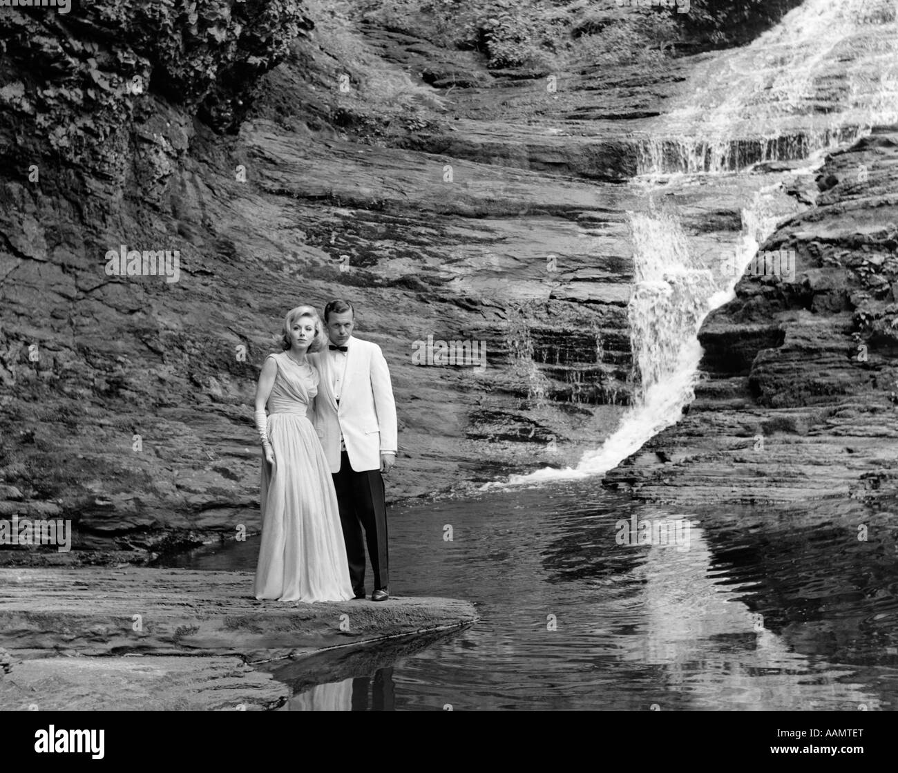 1970s COUPLE IN FORMAL DRESS STANDING ON ROCK AT SIDE OF CREEK WITH WATERFALL COMING DOWN BEHIND THEM Stock Photo