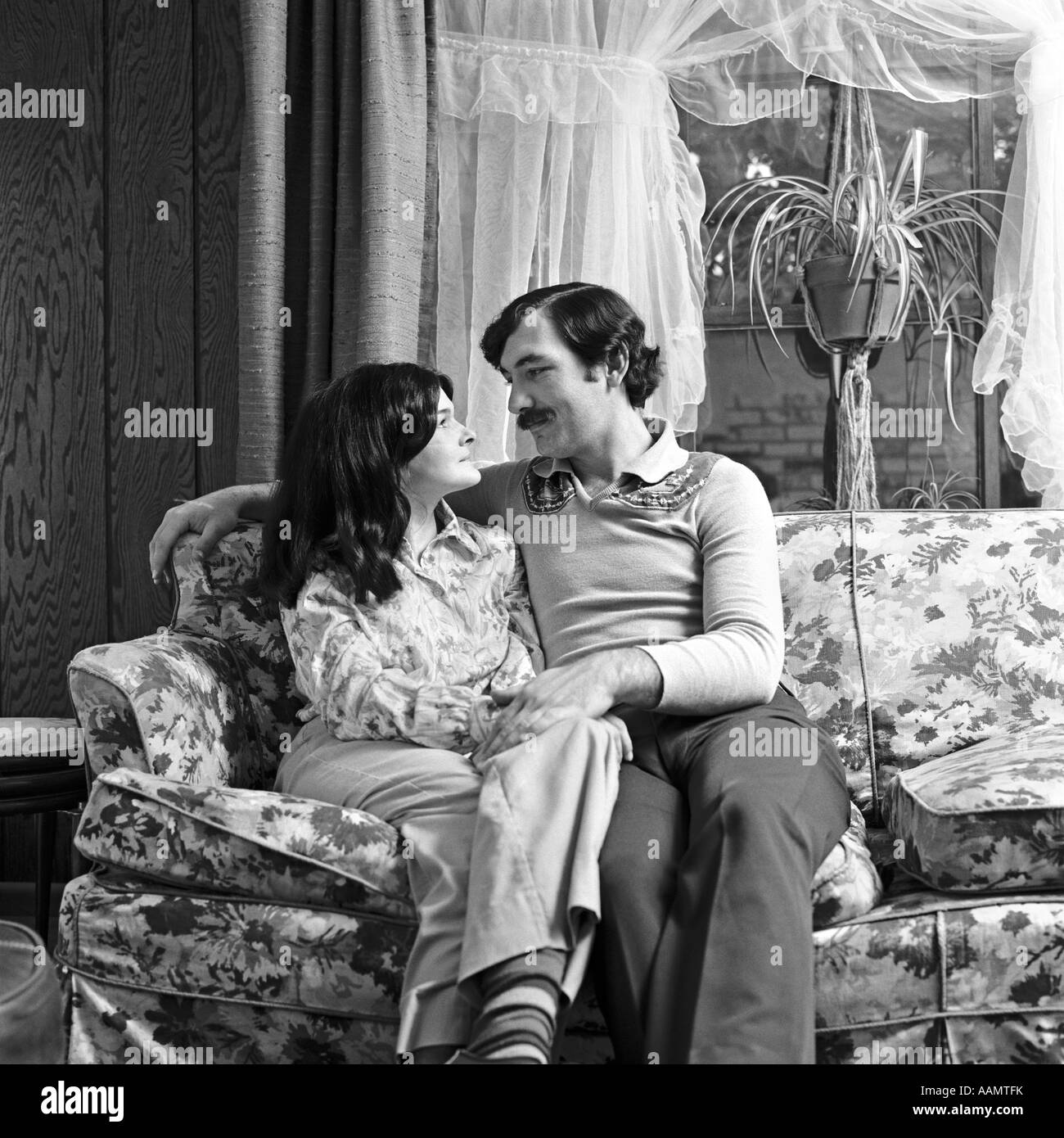 1970s COUPLE HUSBAND WIFE SITTING TOGETHER ON COUCH HUGGING HOLDING HANDS ROMANTIC Stock Photo