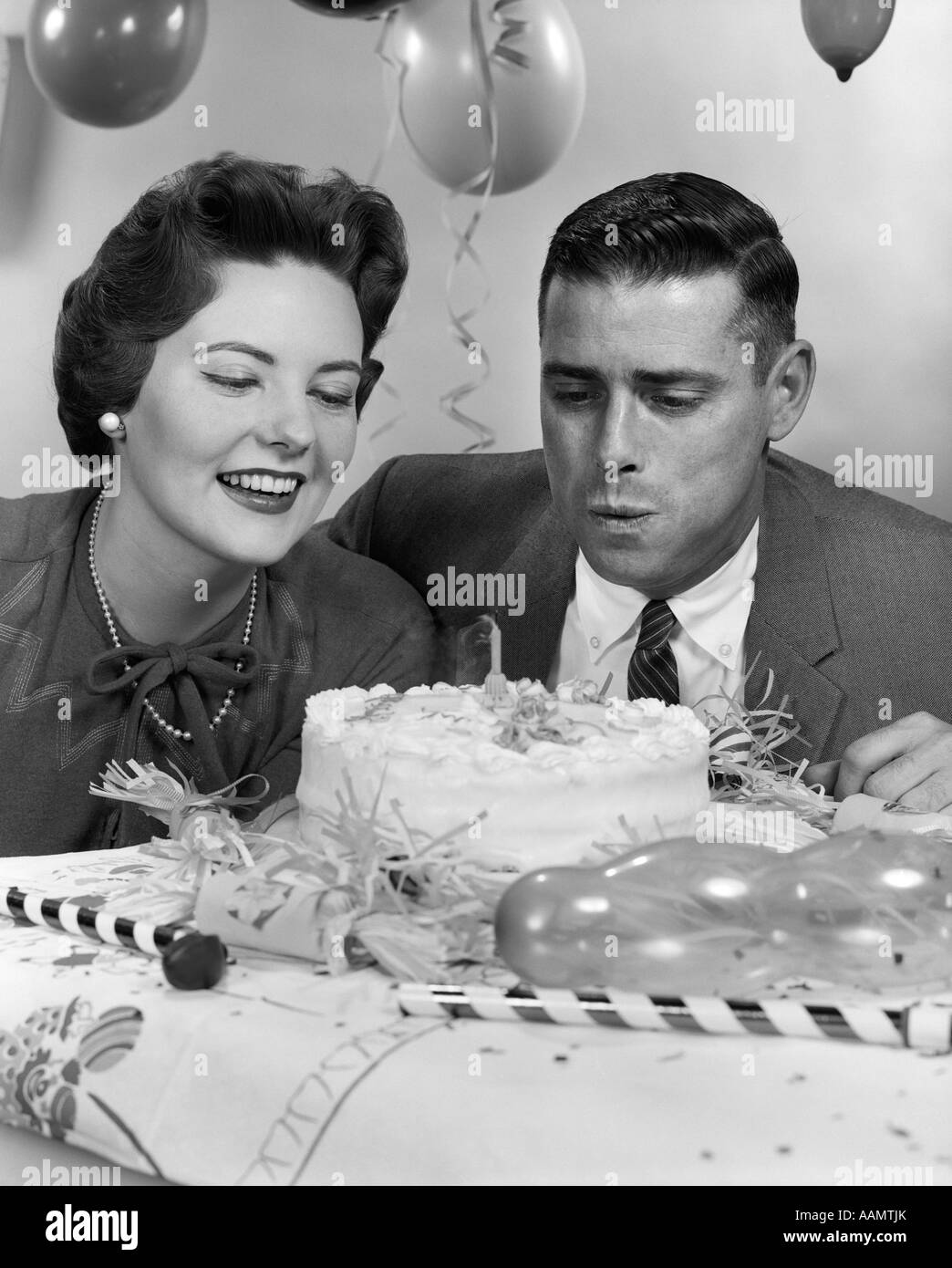 1950s 1960s COUPLE WITH BIRTHDAY ANNIVERSARY CAKE MAN BLOWING OUT CANDLE Stock Photo