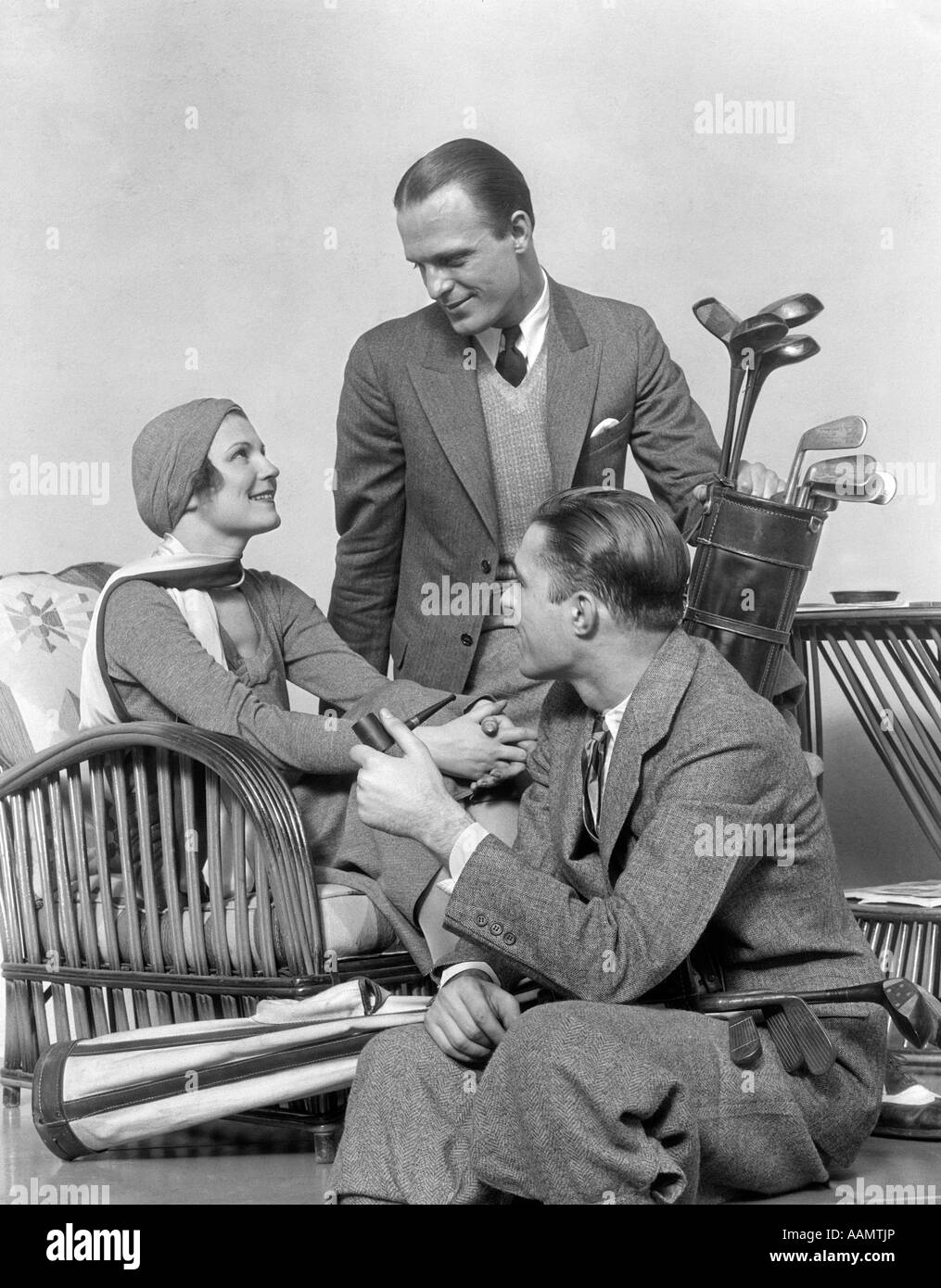 1930s TWO MEN ONE WOMAN GOLF CLUBS BAG SMILING TALKING SITTING BAMBOO CHAIR MAN SMOKING PIPE Stock Photo