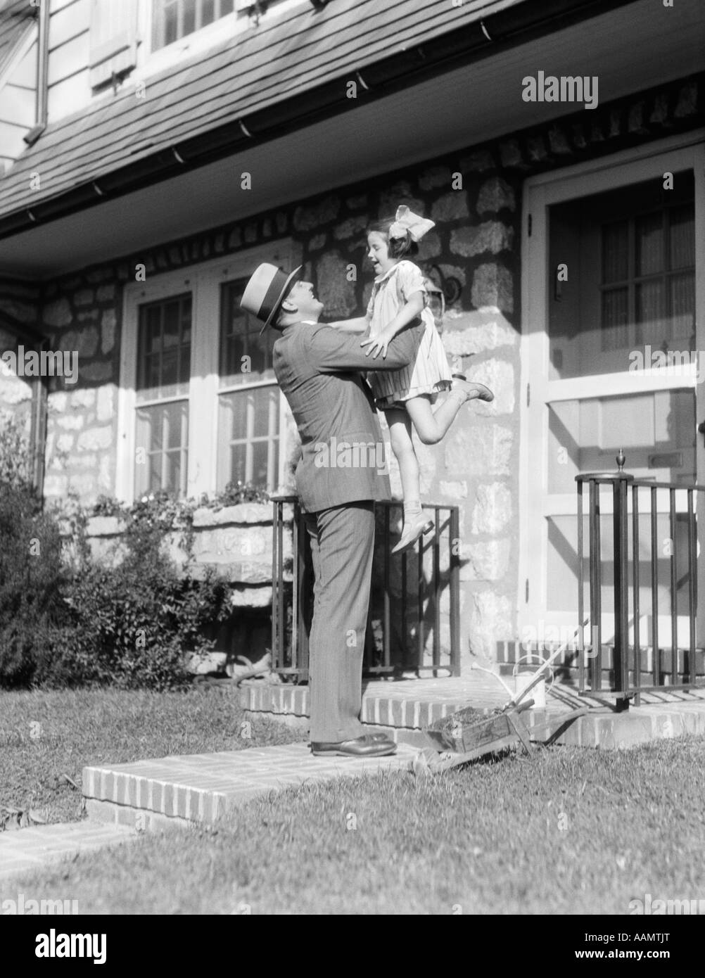 1930s RETURNING FATHER LIFTING UP LAUGHING DAUGHTER IN FRONT OF STONE SUBURBAN HOUSE Stock Photo