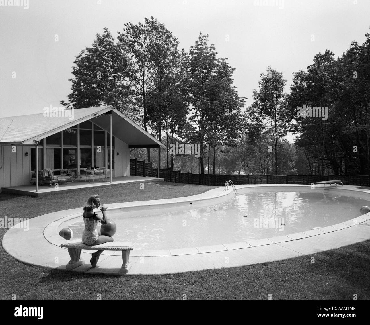 1960s BLOND IN BATHING SUIT SITTING ON STONE BENCH AT END OF KIDNEY-SHAPED POOL Stock Photo