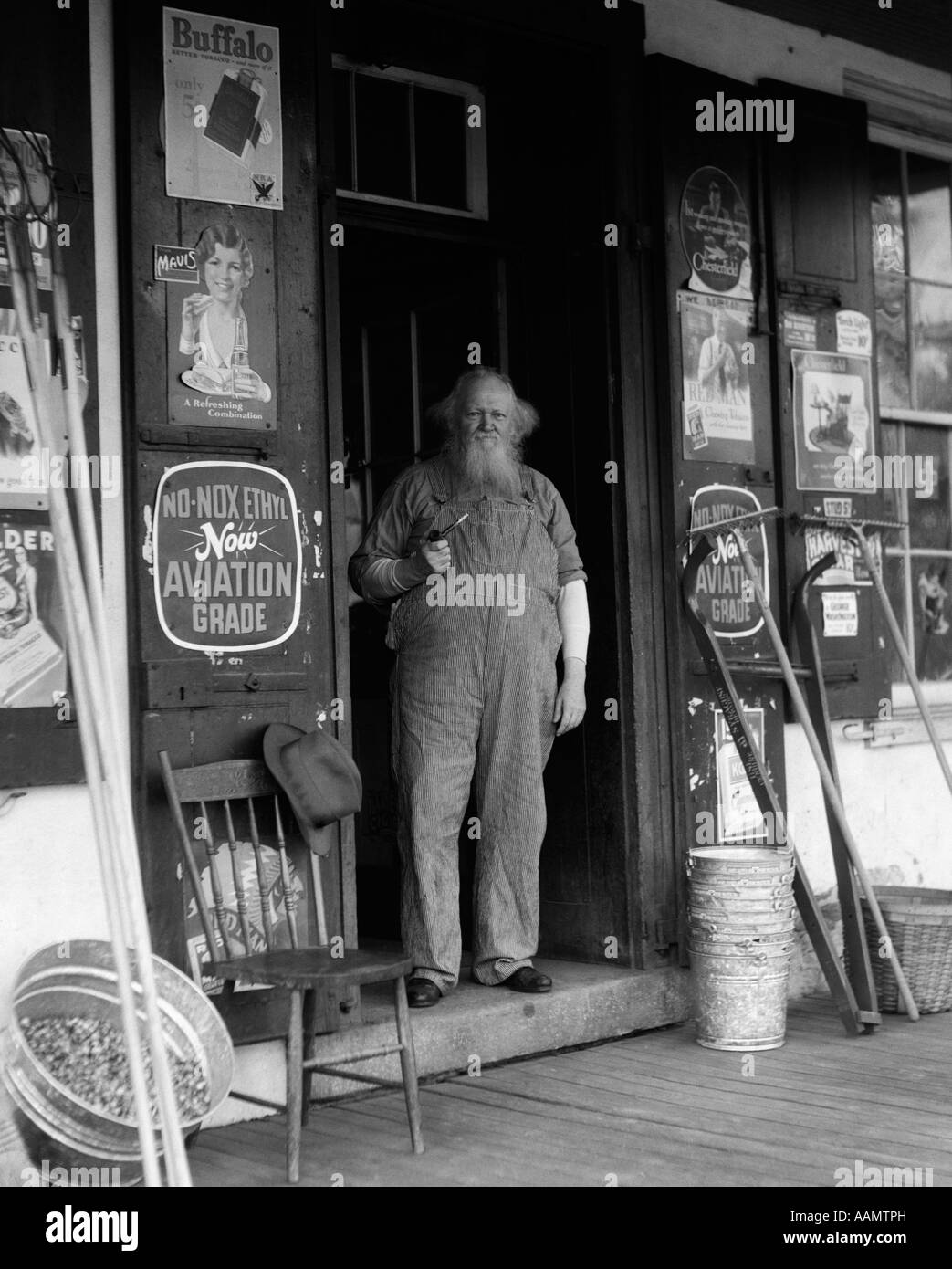 1930s ELDERLY BEARDED MAN IN OVERALLS SMOKING PIPE STANDING IN DOORWAY OF GENERAL STORE Stock Photo