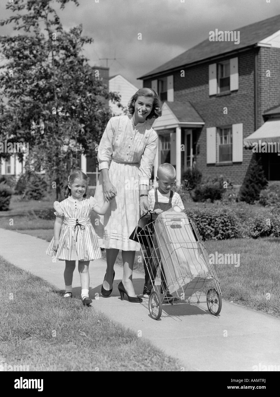 1950s MOTHER LOOKING AT CAMERA SMILING WALKING DOWN SUBURBAN SIDEWALK HOLDING DAUGHTER'S HAND HELPING SON PUSHING GROCERY CART Stock Photo