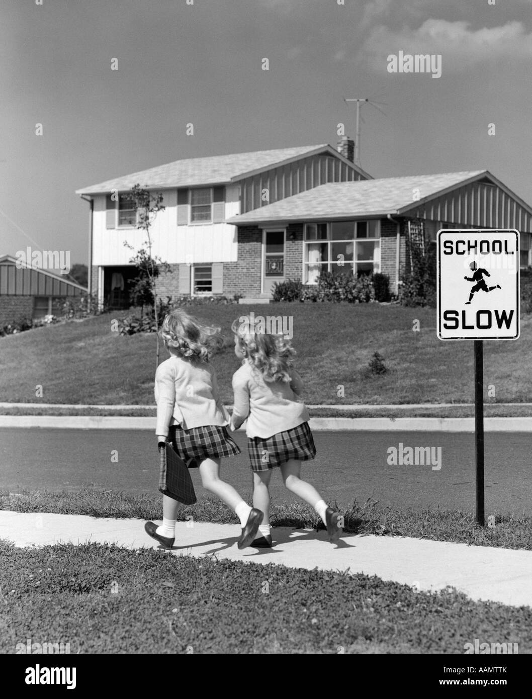 1950s 3 4 BACK VIEW OF TWIN GIRLS IN PLAID SKIRTS & CARDIGANS HOLDING BOOK BAGS RUNNING PAST SCHOOL SLOW SIGN Stock Photo