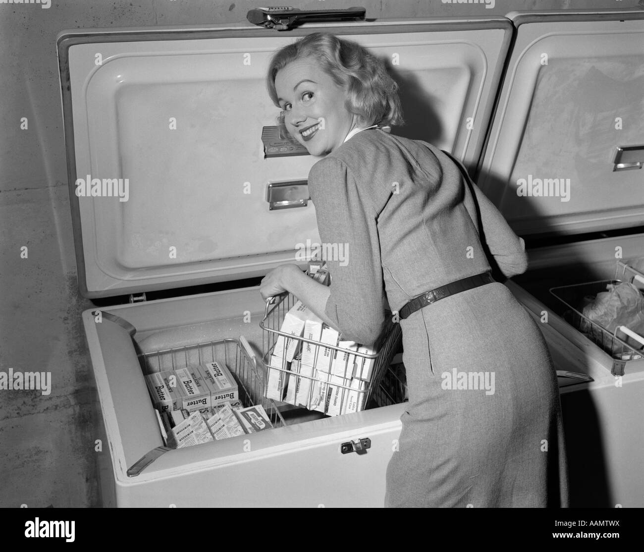 1950s BLOND WOMAN LIFTING WIRE BASKET FOOD ITEMS FROM A DEEP FREEZER LOOKING AT CAMERA OVER HER SHOULDER Stock Photo