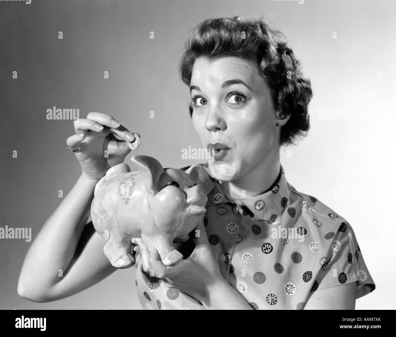 1950s WOMAN LOOKING AT CAMERA DROPPING MONEY IN PIGGY BANK Stock Photo