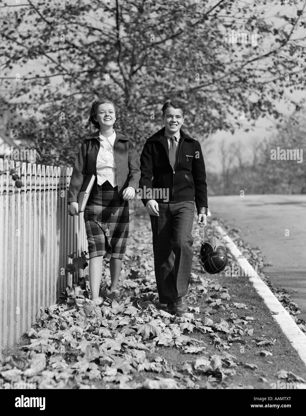 1930s TEEN COUPLE WALKING ON SIDEWALK IN FALL GIRL CARRYING SCHOOLBOOKS