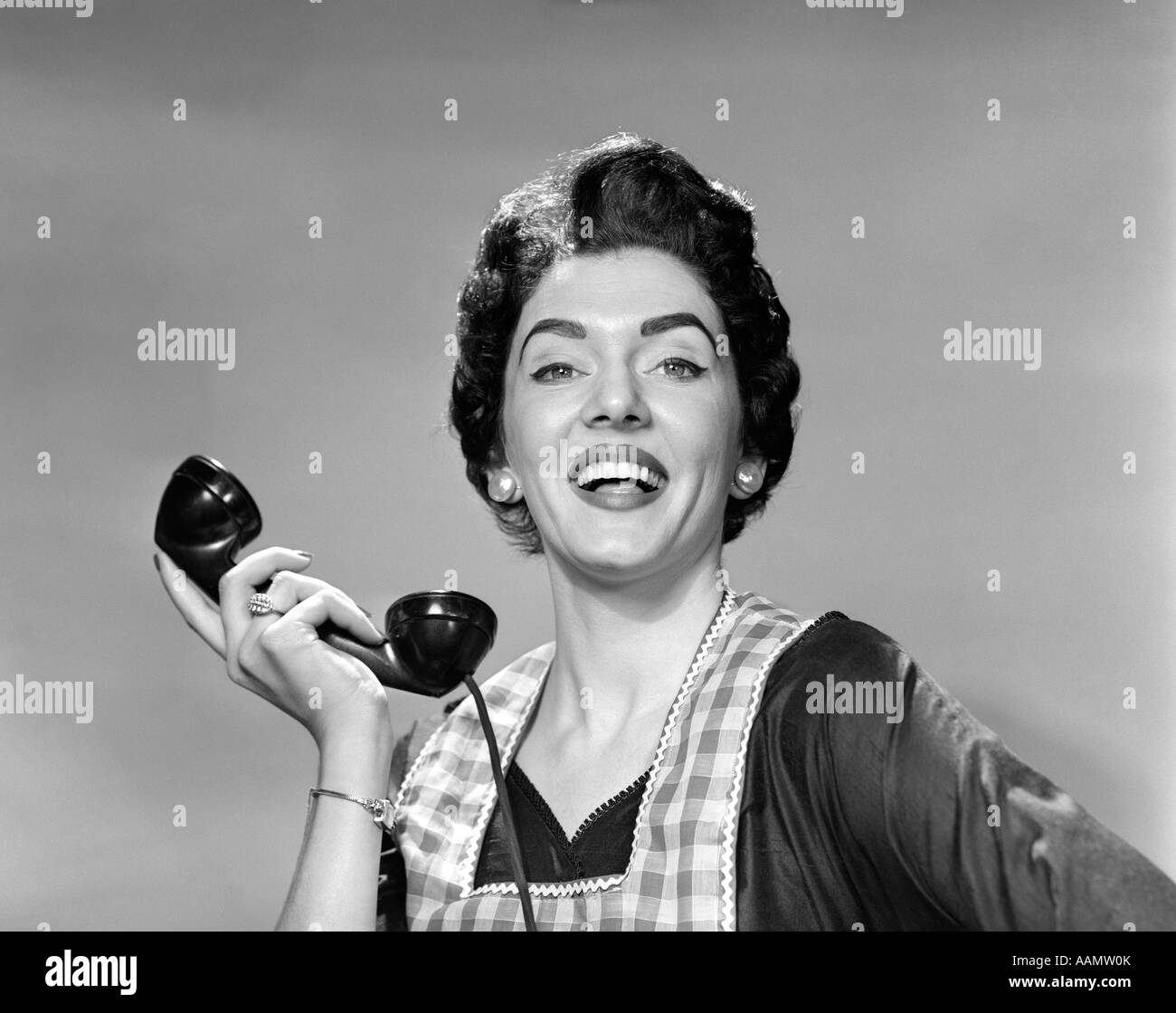 1950s WOMAN IN APRON HOLDING TELEPHONE RECEIVER IN HAND AWAY FROM EAR LOOKING AT CAMERA SMILING Stock Photo