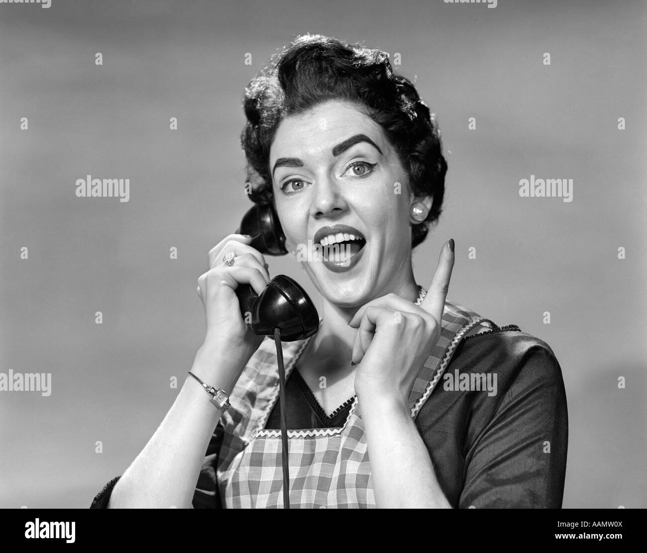 1950s WOMAN IN APRON TALKING ON TELEPHONE POINTING FINGER LOOKING AT CAMERA Stock Photo