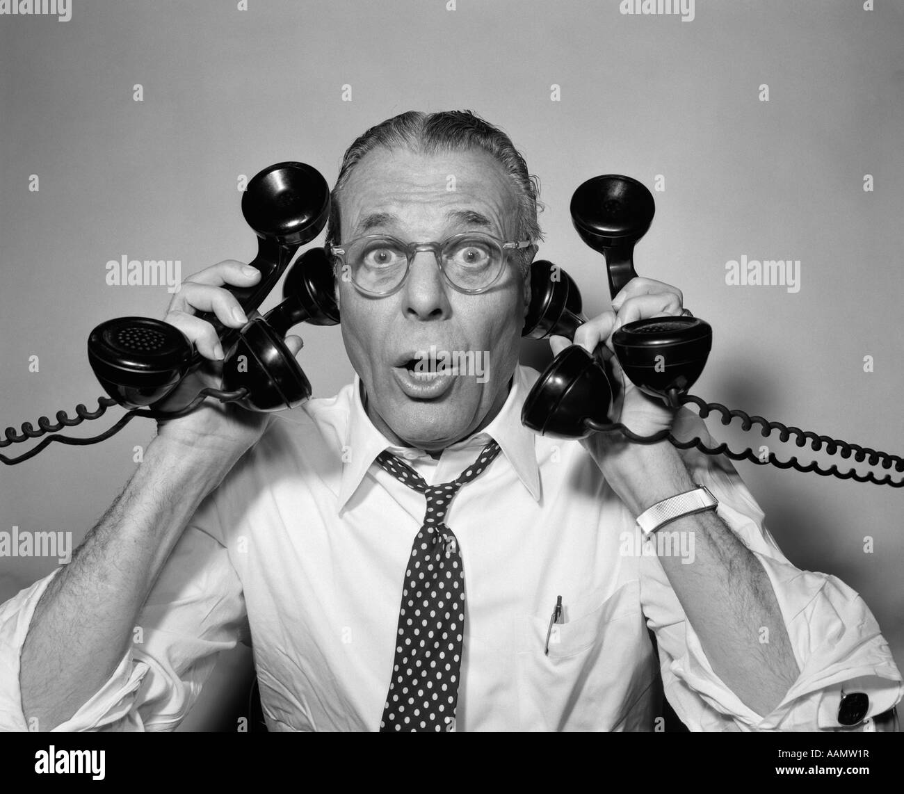 1950s MAN WITH GLASSES HOLDING FOUR BLACK TELEPHONES LOOKING AT CAMERA WITH FUNNY FACIAL EXPRESSION Stock Photo