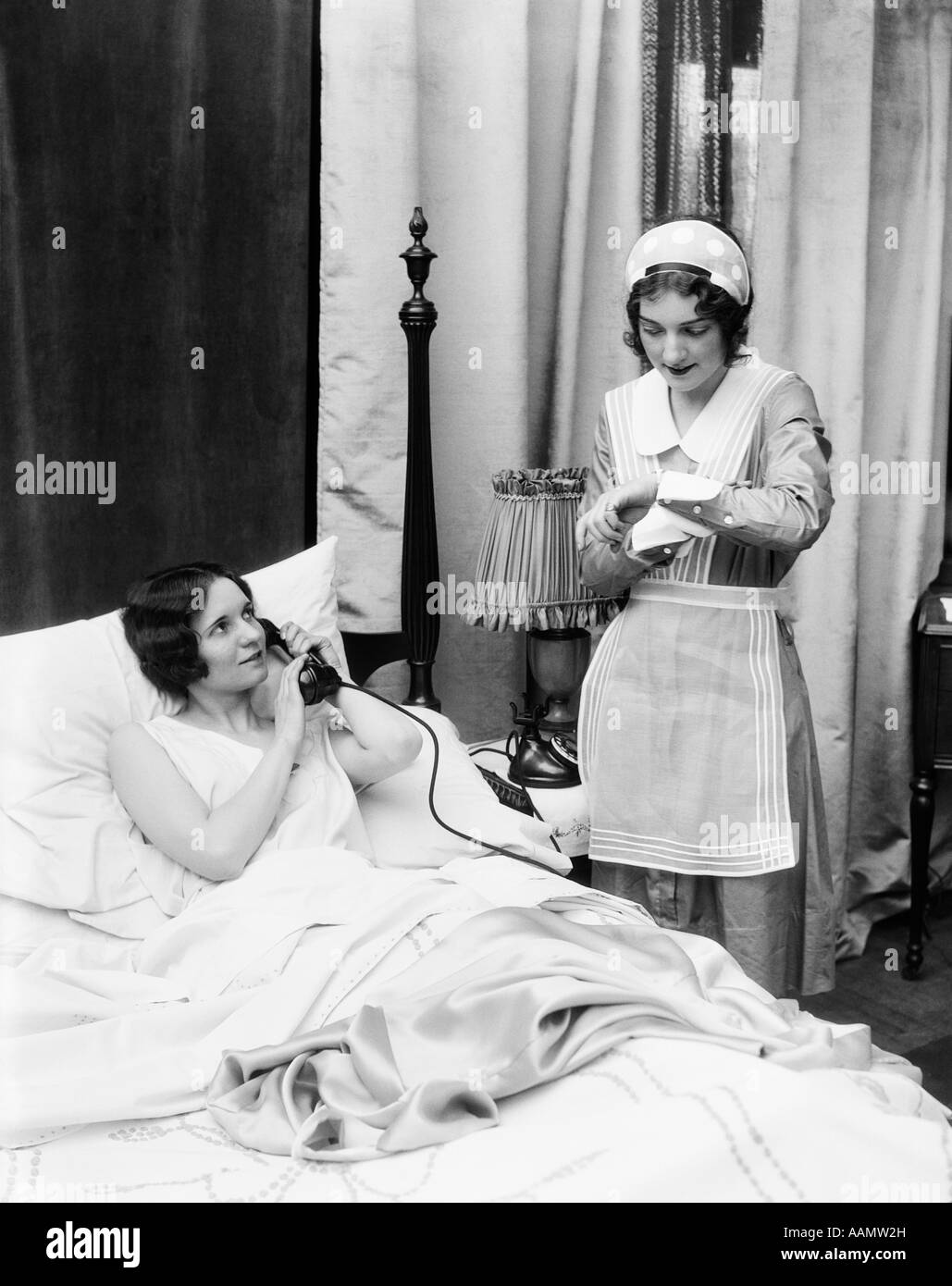 1920s 1930s TWO WOMEN IN BEDROOM MAID LOOKING AT WRIST WATCH OTHER WOMAN IN BED TALKING ON PHONE HAND HELD OVER MOUTHPIECE Stock Photo