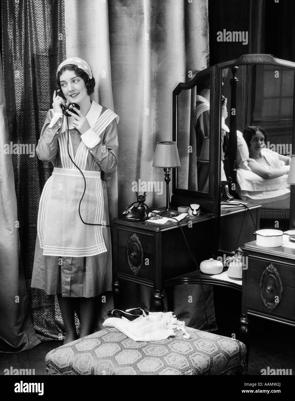 1920s 1930s MAID IN UNIFORM TALKS ON TELEPHONE IN FRONT OF VANITY DRESSING TABLE OTHER WOMAN IS SEEN AS REFLECTION IN MIRROR Stock Photo