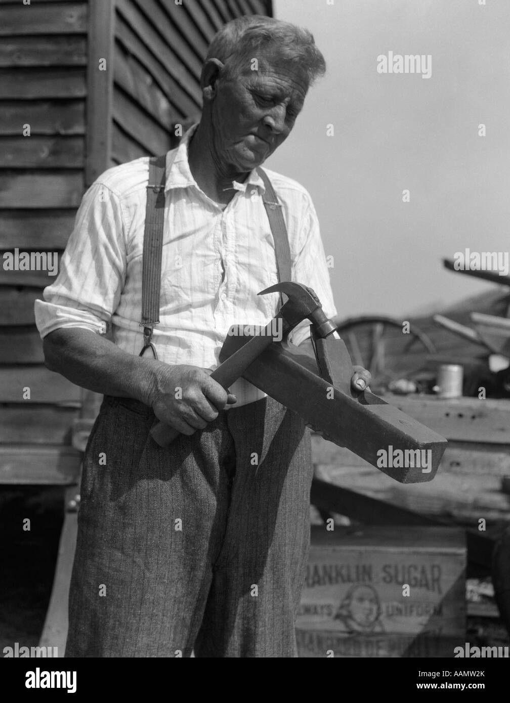 1920s OLD WRINKLED FACE MAN STANDING IN FRONT OF CLAPBOARD BUILDING HOLDING A PLANE AND HAMMER Stock Photo