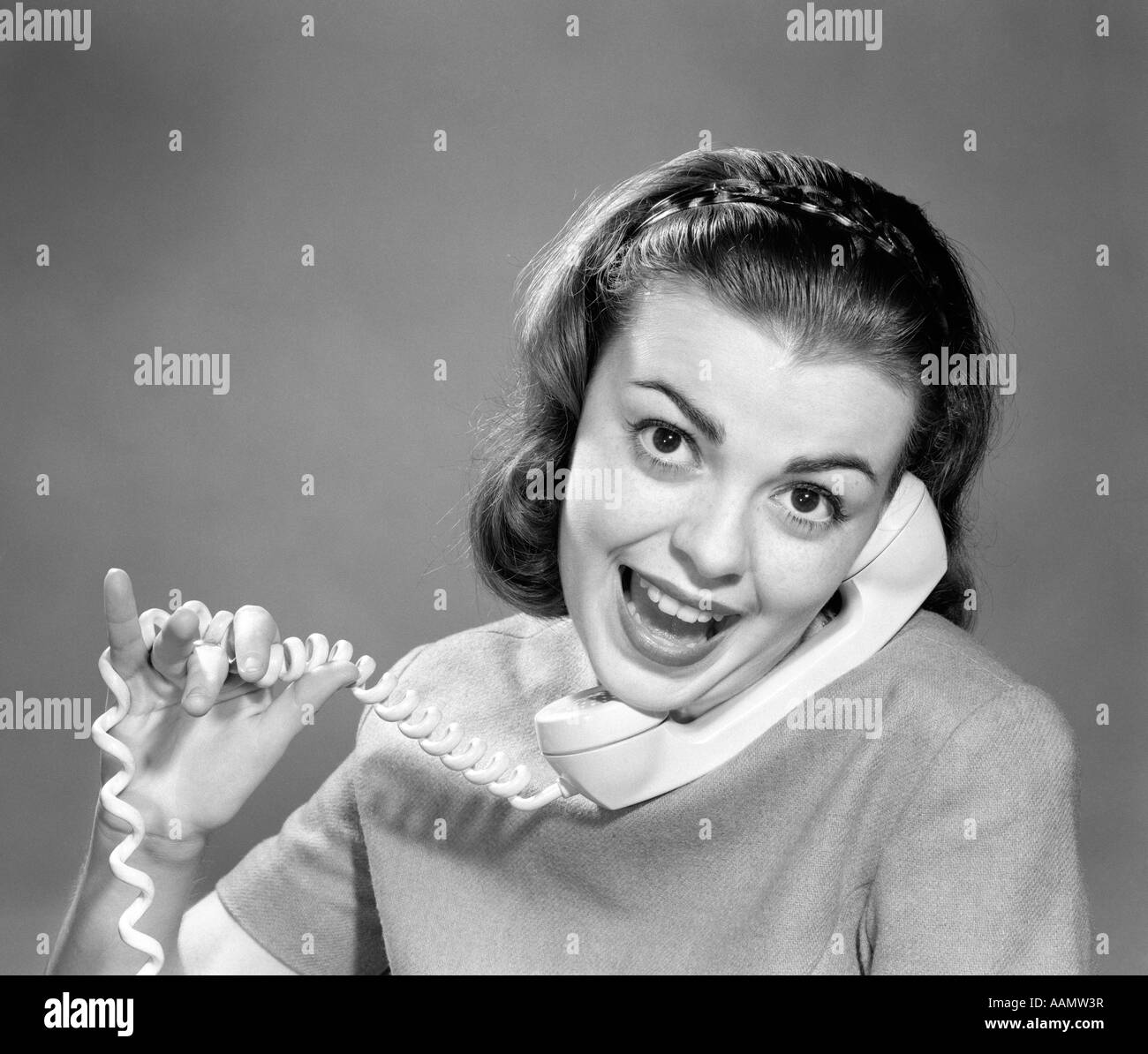 1950s 1960s YOUNG WOMAN TALKING ON TELEPHONE CORD WRAPPED IN FINGERS LOOKING AT CAMERA Stock Photo