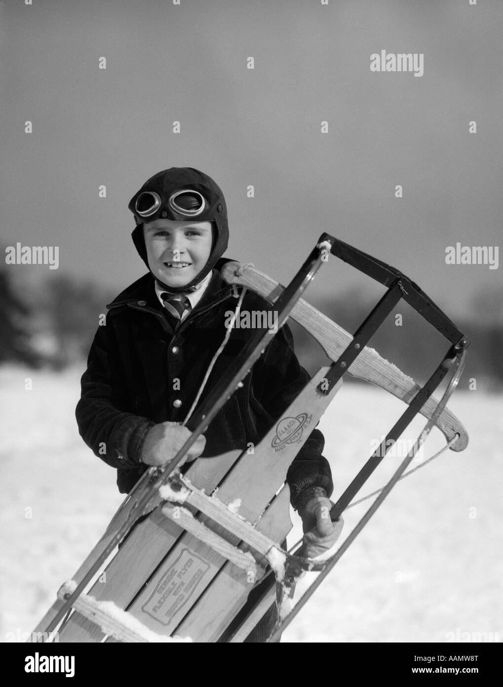 1920s 1930s SMILING BOY WEARING AVIATOR GOGGLES LEATHER FLYING HELMET HOLDING SLED STANDING IN SNOW FIELD LOOKING AT CAMERA Stock Photo