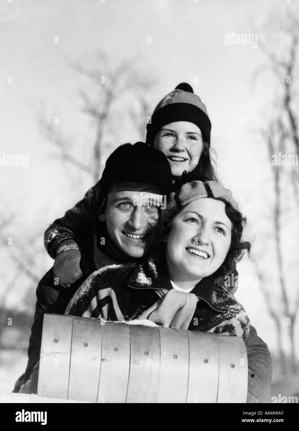 1930s MOTHER FATHER CHILD LYING ON TOBOGGAN FACING CAMERA SMILING HAT SCARF HOOD Stock Photo