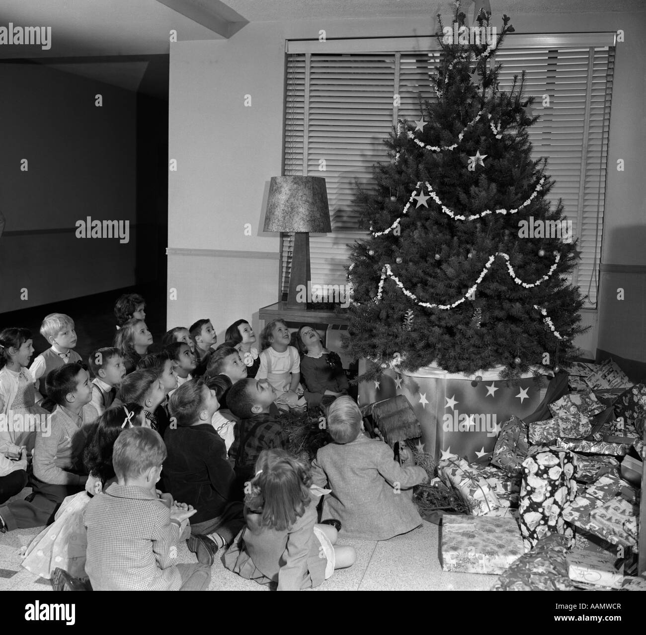 1950s SEATED GROUP OF 20 CHILDREN LOOKING UP AT CHRISTMAS TREE DECORATED WITH POPCORN AND STARS Stock Photo