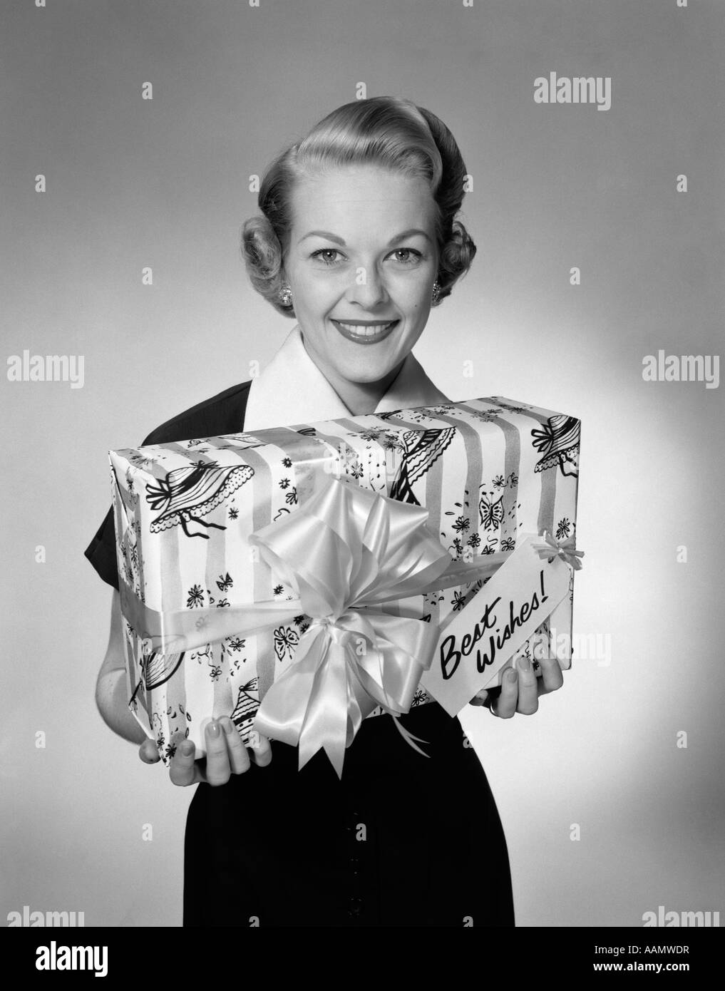 1950s WOMAN HOLDING WRAPPED PRESENT WITH BEST WISHES TAG LOOKING AT CAMERA Stock Photo
