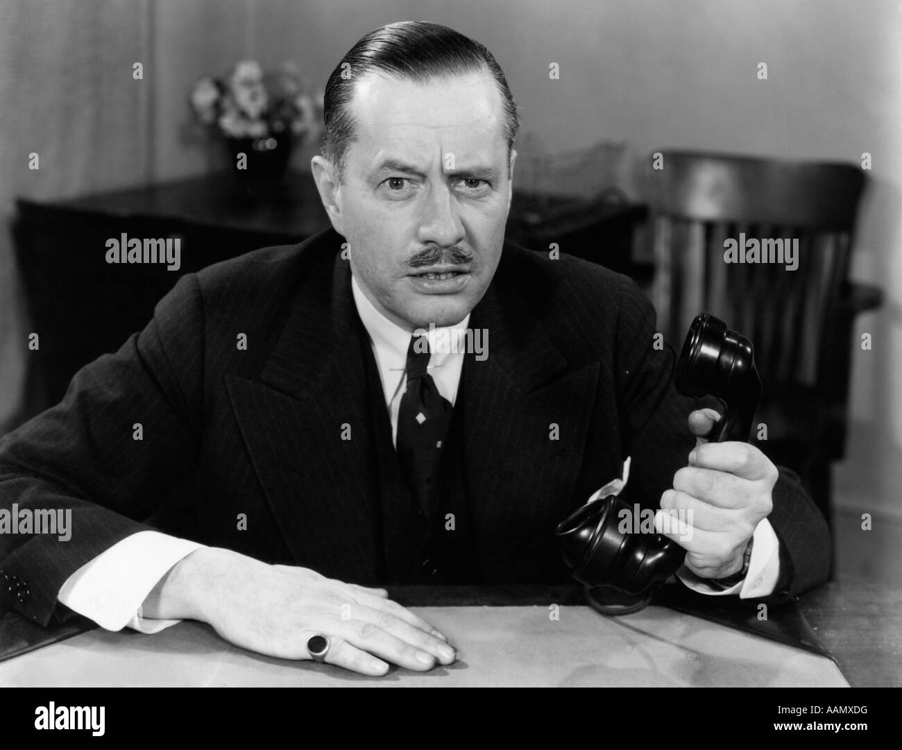 1930s 1940s PORTRAIT OF BUSINESS MAN SITTING AT OFFICE DESK HOLDING PHONE IN ONE HAND SERIOUS FACIAL EXPRESSION MOUSTACHE Stock Photo