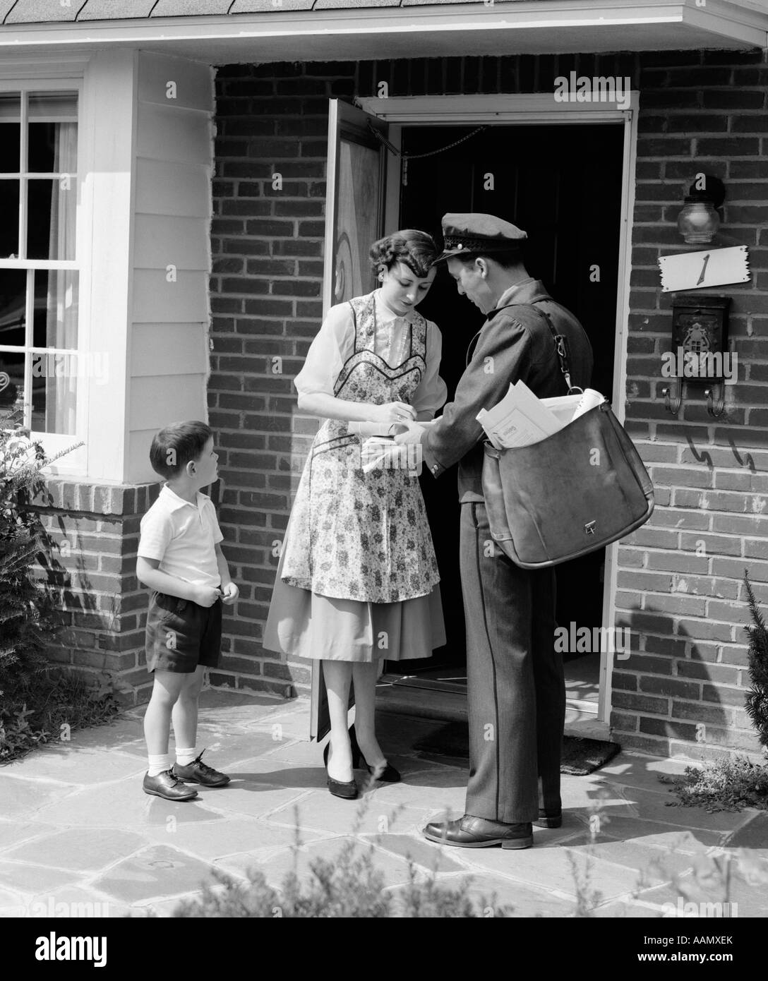 1950s SUBURBAN MOM AT HOME FRONT DOOR WITH SON WATCHING RECEIVING PACKAGE FROM MAILMAN Stock Photo