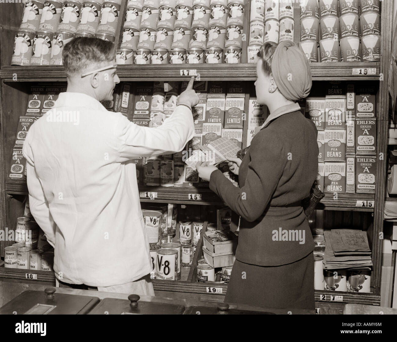 1940s BACK VIEW OF GROCER POINTING OUT PRICES OF CANNED GOODS TO WOMAN HOLDING RATION BOOK Stock Photo