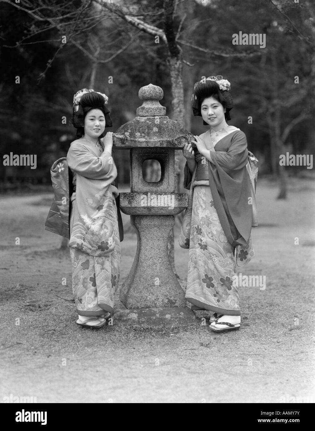 1930s TWO JAPANESE GEISHA GIRLS IN NATIVE COSTUME KIMONO STANDING BY STONE LANTERN KOBE JAPAN Stock Photo