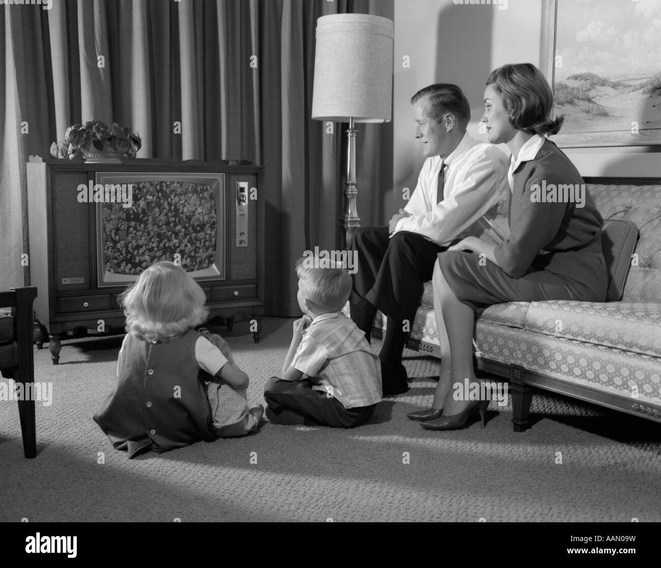 1960s FAMILY FOUR IN LIVING ROOM WATCHING TELEVISION MOM DAD ON COUCH BOY GIRL SIT ON FLOOR Stock Photo
