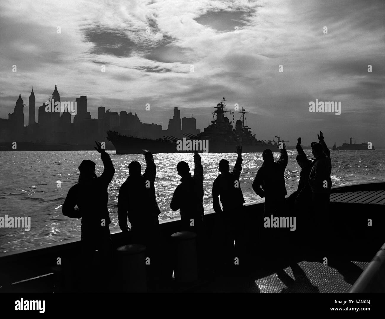 1940s 1950s SILHOUETTED SAILORS DECK OF SHIP WAVING SALUTE TO PASSING USN BATTLESHIP AT NIGHT AGAINST NEW YORK CITY SKYLINE Stock Photo